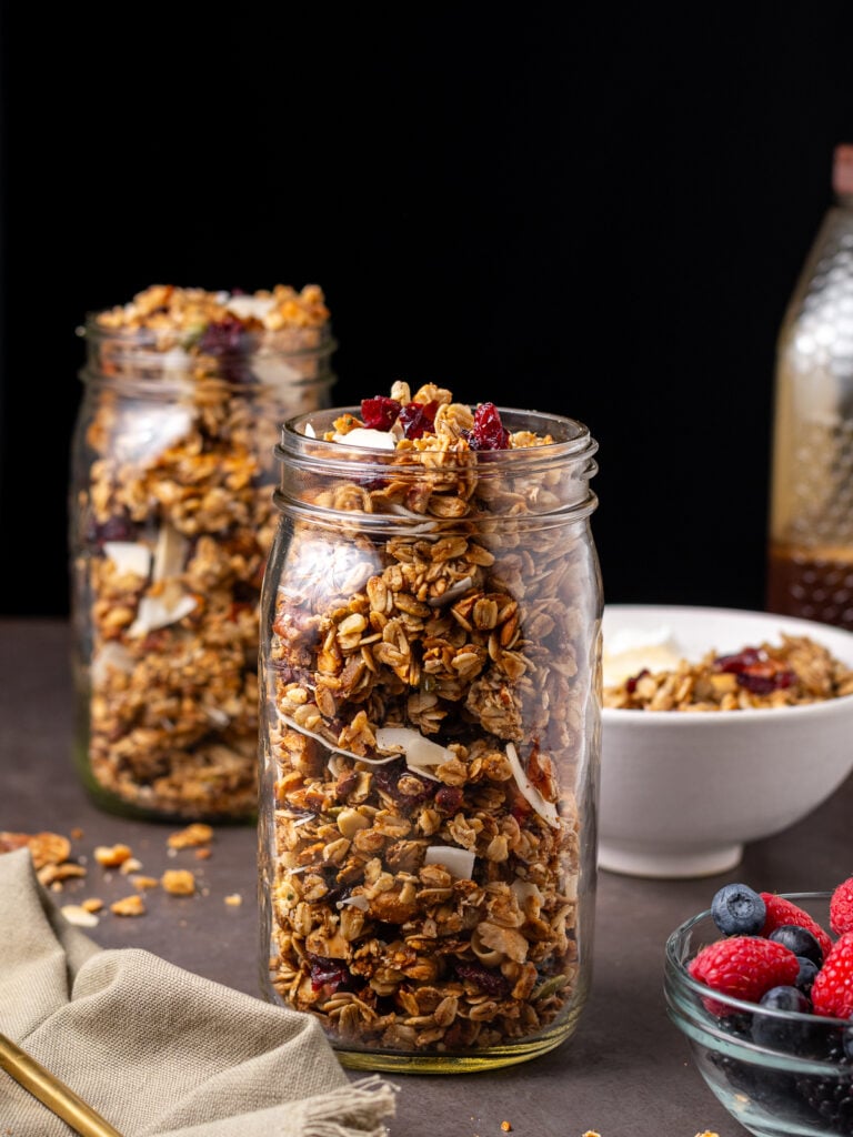 Homemade cinnamon granola in glass mason jars with crispy clusters, walnut, and dried fruit served alongside a bowl of berries.