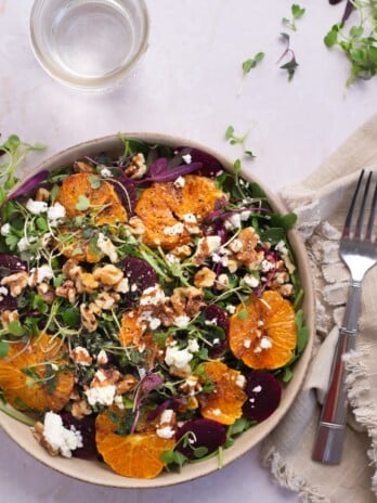 Overhead view of a bowl of arugula salad with oranges, goat cheese, walnuts, pickled beets and microgreens with a napkin, fork and small glass of water