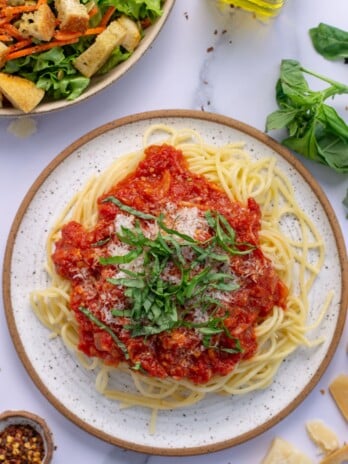 Spaghetti noodles and spicy arrabiata sauce topped with parmesan and basil on a white plate, big bowl of salad on the side