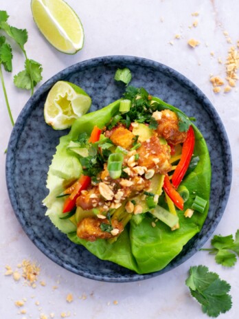 Three lettuce wraps filled crispy tofu, peanut sauce, bell peppers, avocado, cucumber, carrots, fresh herbs and peanuts. Displayed with lime slices, crushed peanuts and cilantro stems.