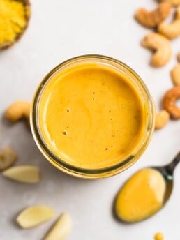 Spicy cashew dressing in a mason jar with a spoonful of dressing, roasted cashews, garlic cloves and a small bowl of nutritional yeast