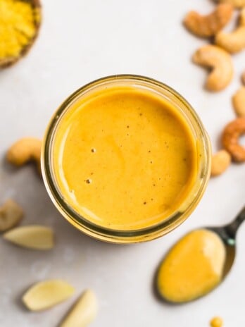 Spicy cashew dressing in a mason jar with a spoonful of dressing, roasted cashews, garlic cloves and a small bowl of nutritional yeast