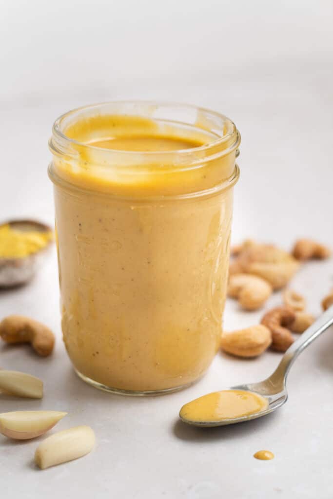 Jar with spicy cashew dressing, spoon with some of the dressing, roasted cashews, small bowl of nutritional yeast and 3 garlic cloves