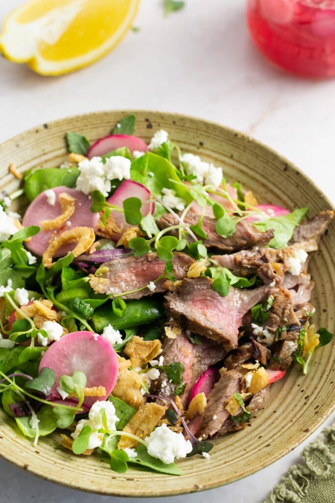 Speckled bowl filled with an arugula salad topped with pickled radishes, crispy onions, medium rare thinly sliced steak, microgreens and goat cheese.