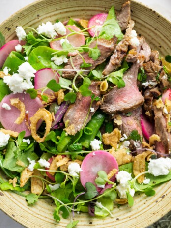 speckled bowl filled with salad containing arugula, butter lettuce, steak, microgreens, radishes, snap peas, crispy onions and goat cheese