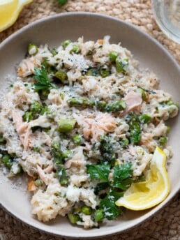 Smoked salmon risotto on a plate with peas, asparagus, fresh herbs and a lemon wedge. Glass of wine on the side.