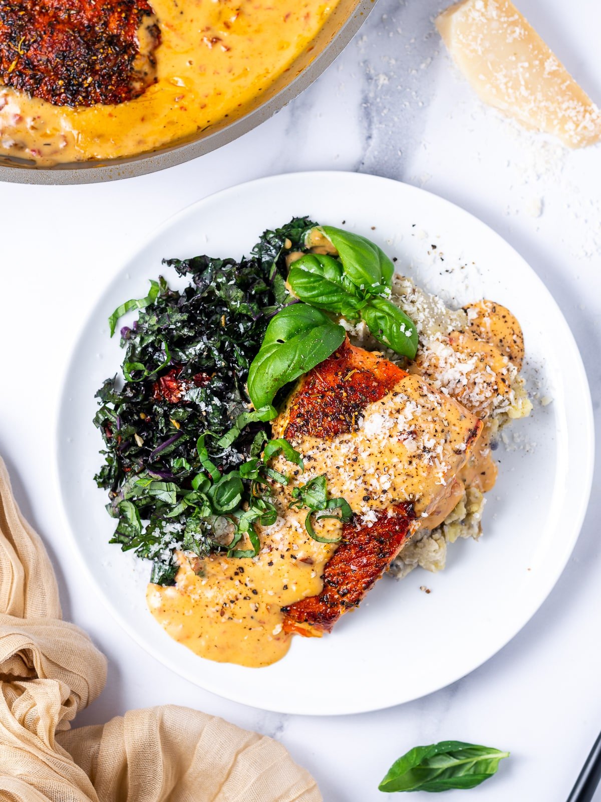 white plate with seasoned salmon, creamy sun-dried tomato sauce, fresh basil, sauteed kale and mashed potatoes