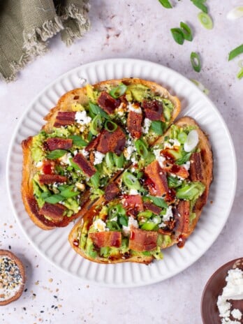 white plate with two pieces of avocado toast topped with bacon and feta