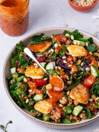 bowl of Greek spinach salad with fried feta, spoonful of tomato dressing, kalamata olives, cucumber and pine nuts
