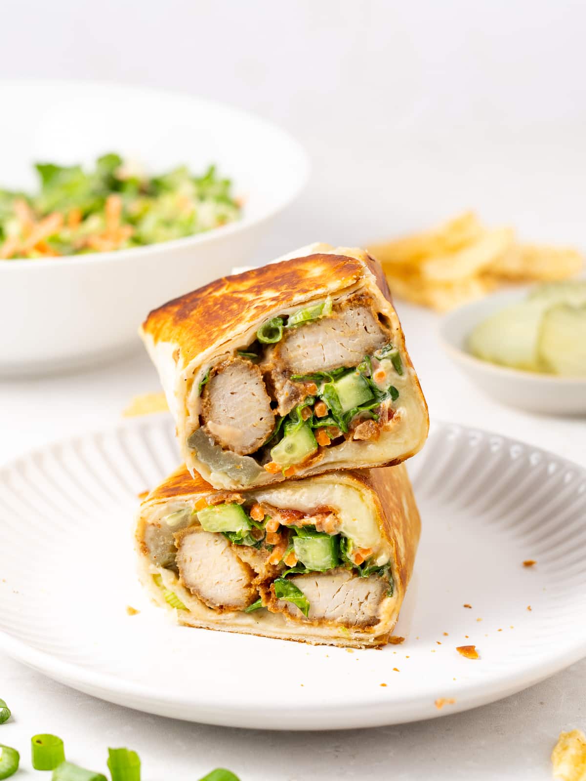 two halves of a honey mustard chicken wrap on a white plate with pickles, salad and potato chips in the background