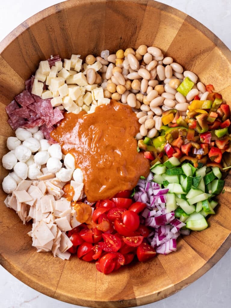 Dense bean salad ingredients of beans, tomatoes, cucumbers, peppers, deli meat, and cheese topped with sun-dried tomato vinaigrette dressing before mixing