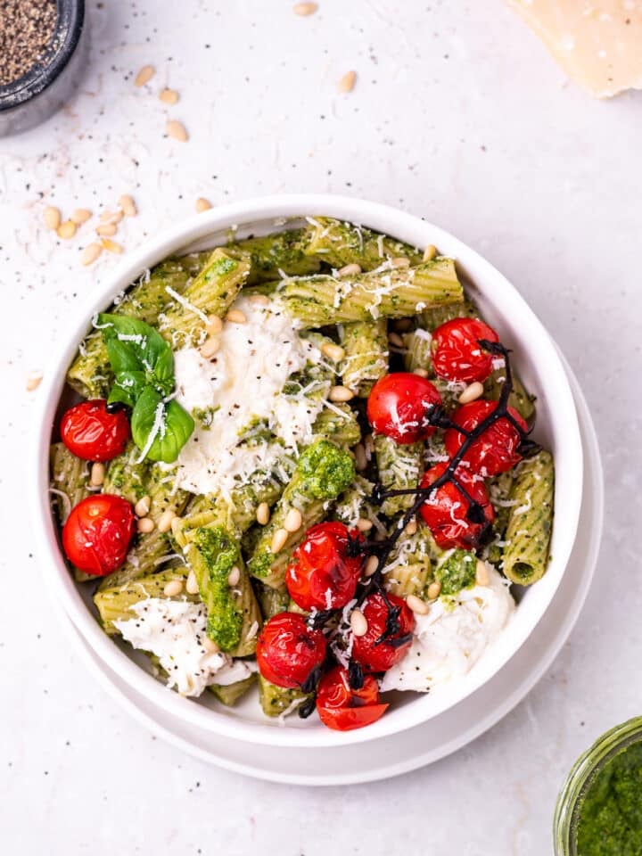white bowl with rigatoni pasta covered in pesto topped with burrata cheese and burst tomatoes