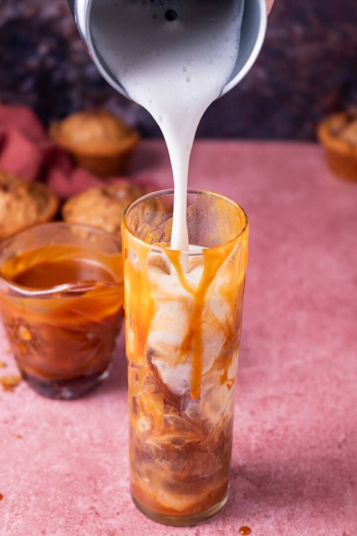 hand pouring frothed oat milk into a glass with ice, espresso and salted caramel drizzle