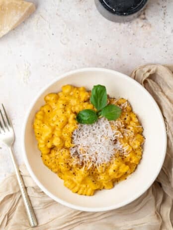 white bowl with butternut squash feta pasta, fork, napkin, pepper grinder and block of parmesan cheese