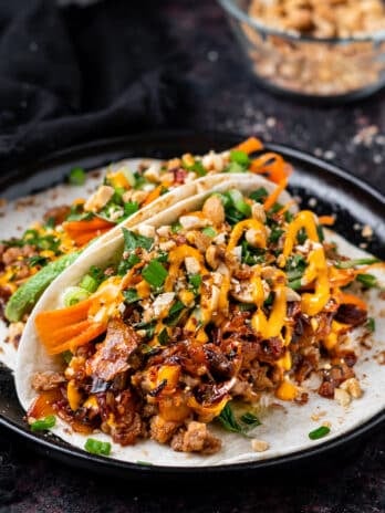 black plate with two tacos filled with Korean chicken, caramelized kimchi, fresh cucumber, avocado and herbs, peanuts and sriracha mayo