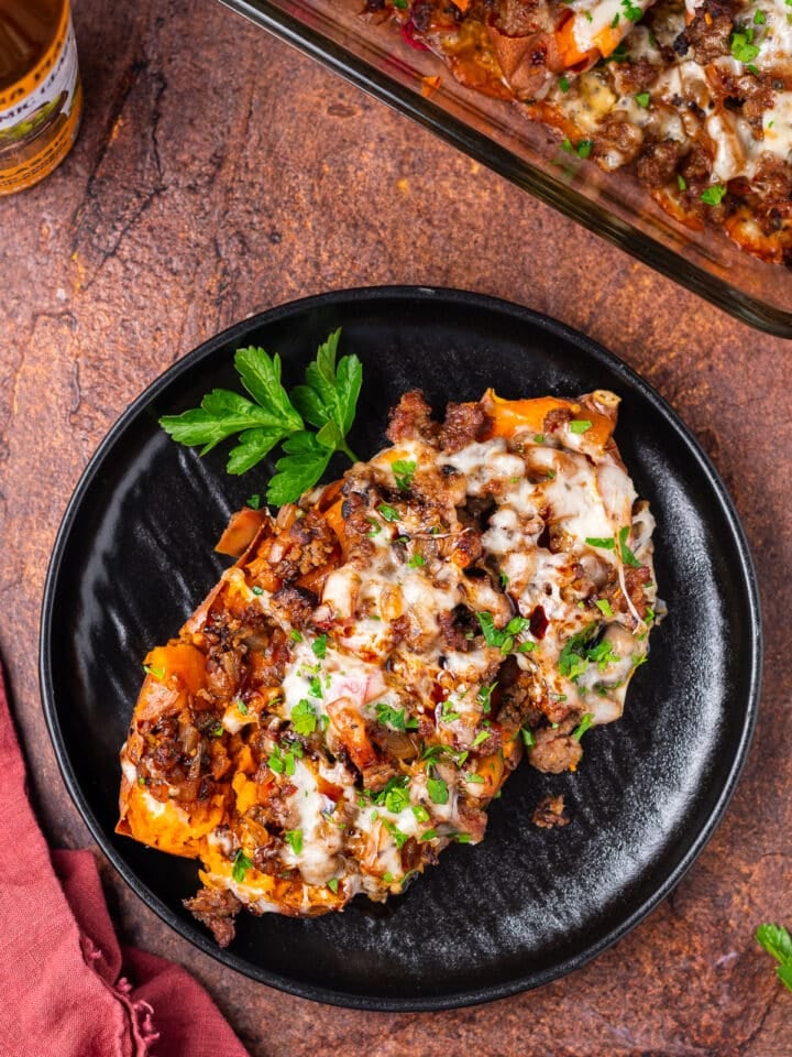 black plate, baked sweet potato with italian sausage, onion, apple and mozzarella cheese, sprig of parsley