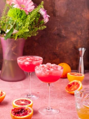 two coupe glasses filled with blood orange juice, seltzer and lemon honey syrup, vase of flowers, halved blood oranges