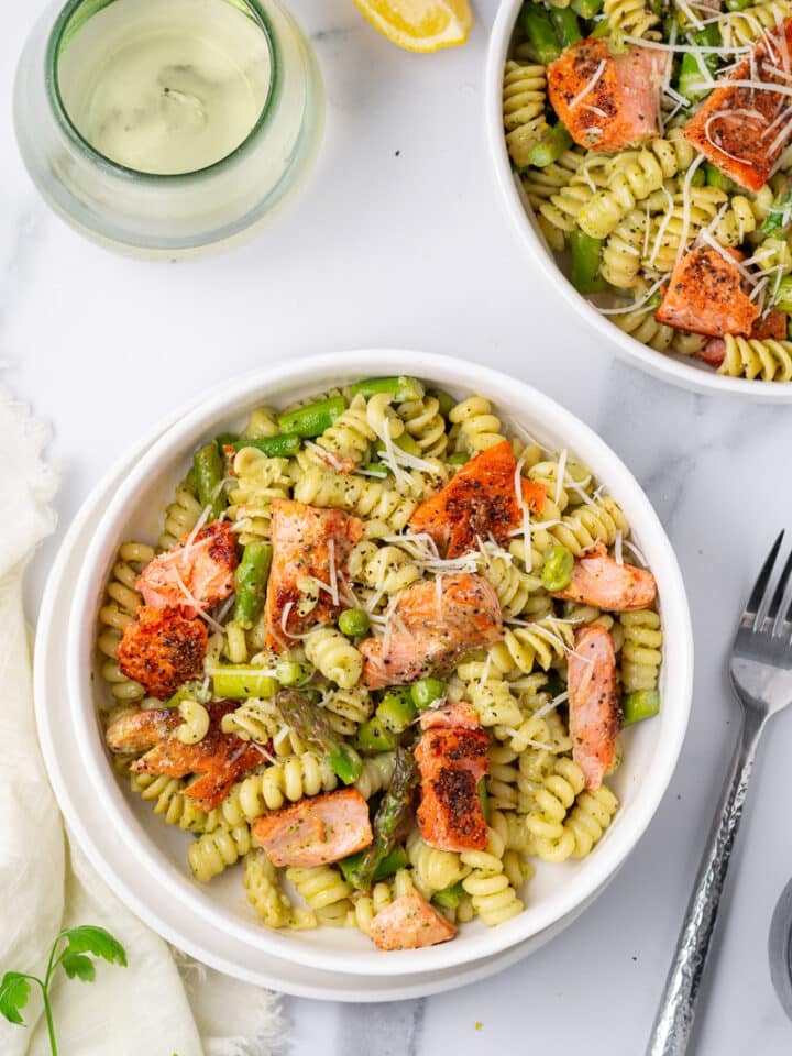two white bowls with pesto pasta, salmon, asparagus and peas