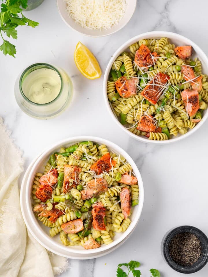 two white bowls of salmon pesto pasta, glass of wine, lemon wedge, black pepper cellar