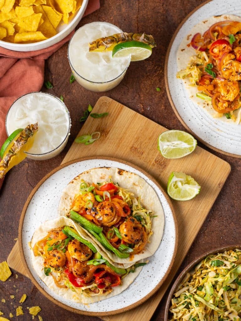 white plate with two tacos with baja shrimp, avocado slices, slaw and fajita veggies, pineapple margarita, lime slices