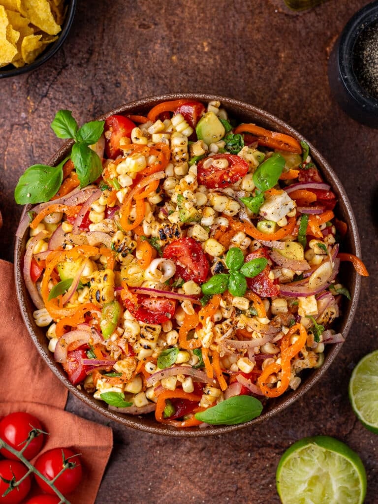 Large bowl filled with charred corn, tomatoes, avocado, red onion, green onions, basil, black pepper, lime juice, honey and olive oil