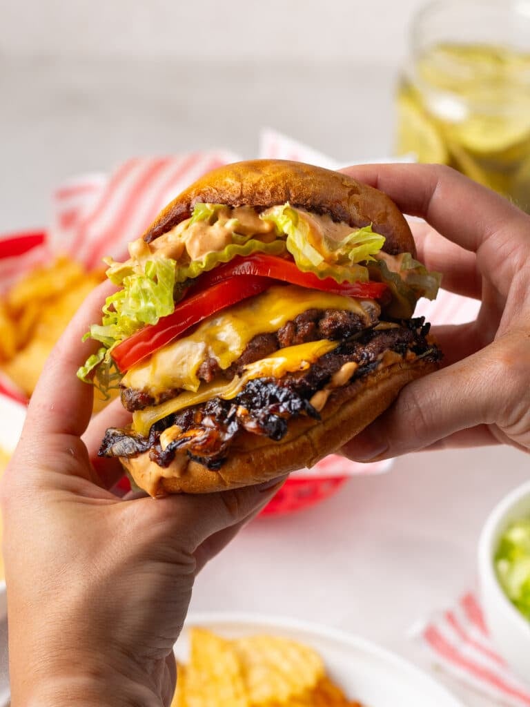 hands holding a smash burger with caramelized onions, tomato, pickles, lettuce and burger sauce