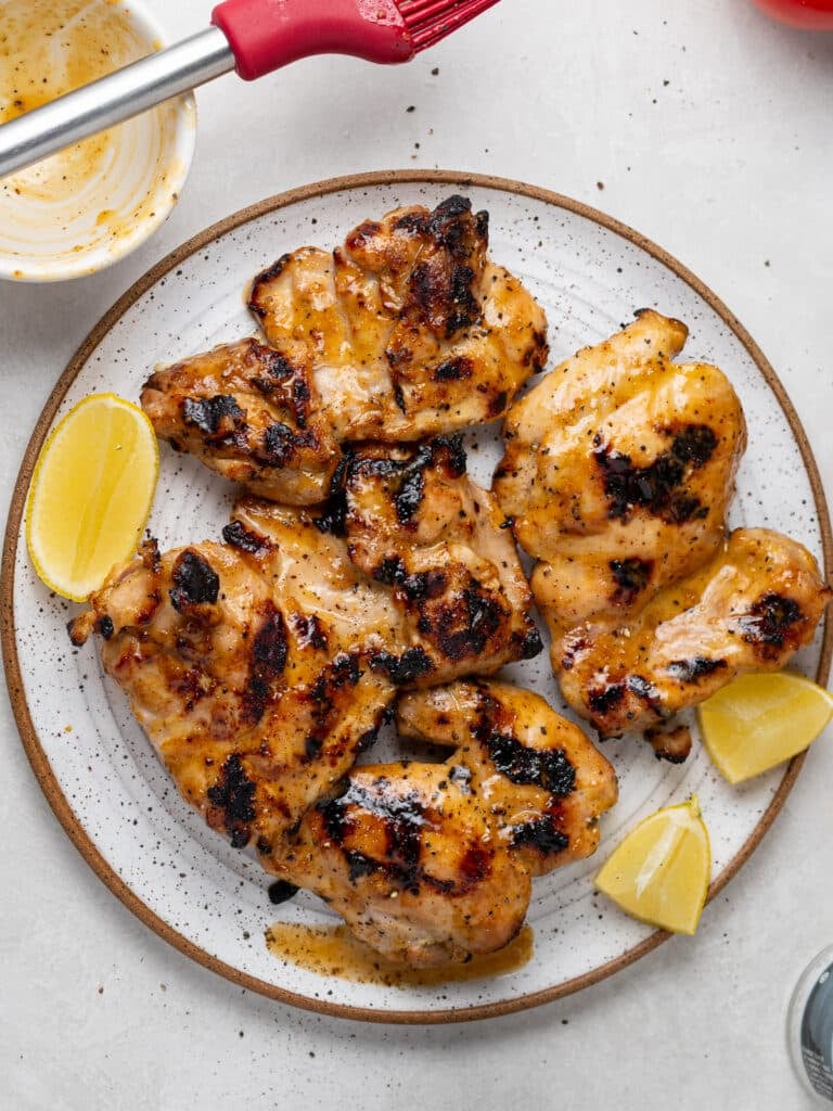perfectly grilled chicken thighs on a white speckled plate with lemon wedges