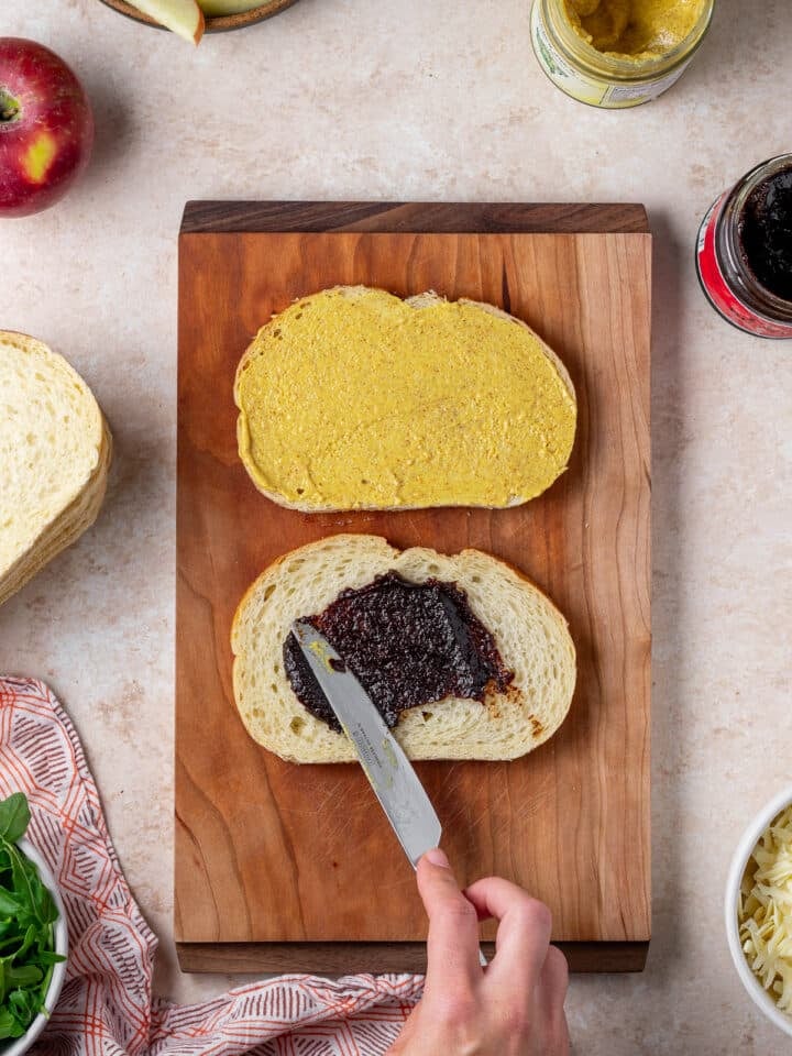 hand using a knife to spread fig butter on one slice of bread and dijon mustard aioli on another slice of bread