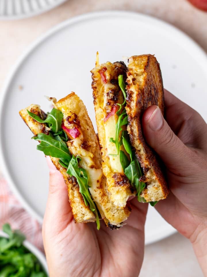 hands holding a grilled cheese sandwich that has Gruyere cheese, bacon, apple, fig jam and arugula