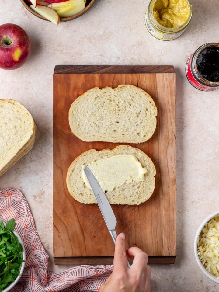 hand using a knife to spread mayo on the outside of two pieces of bread