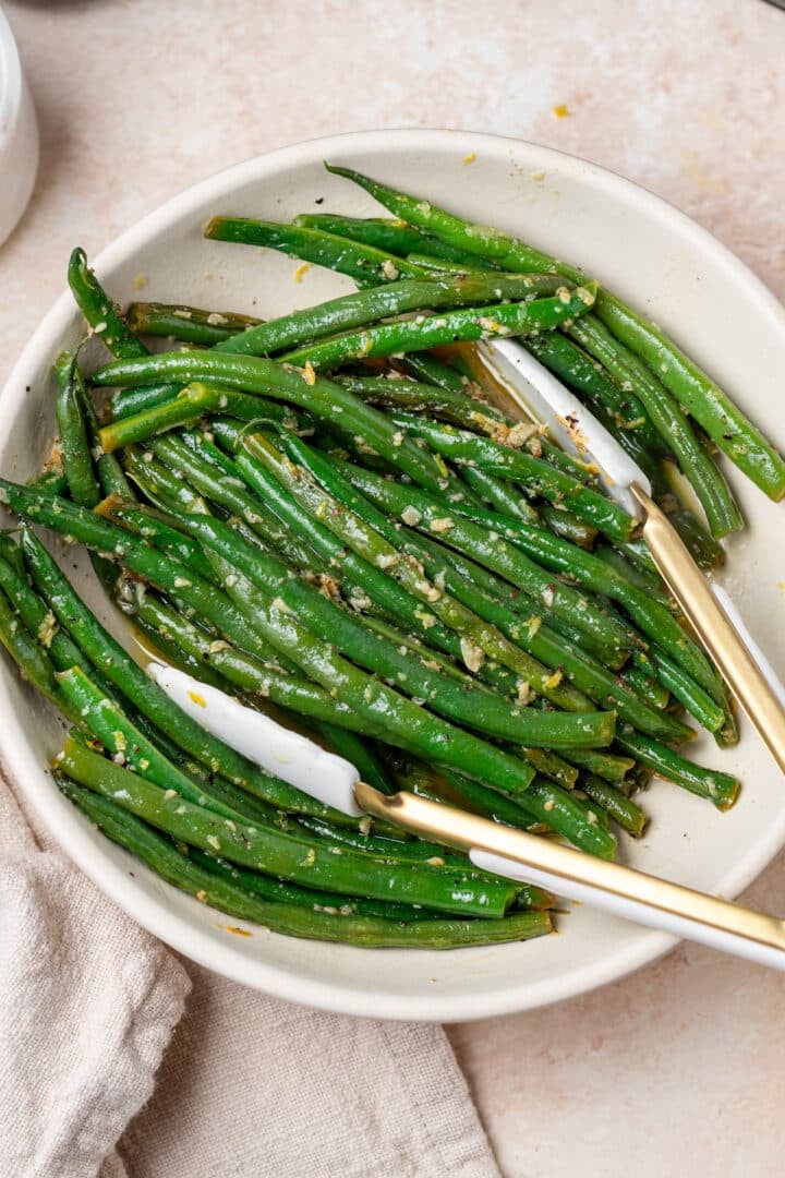 white serving bowl with lemon butter garlic green beans, white and gold tongs