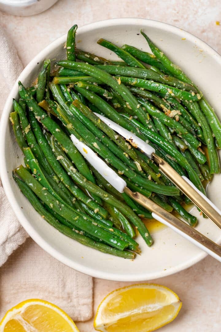 tongs grabbing a portion of haricot verts from a white serving bowl