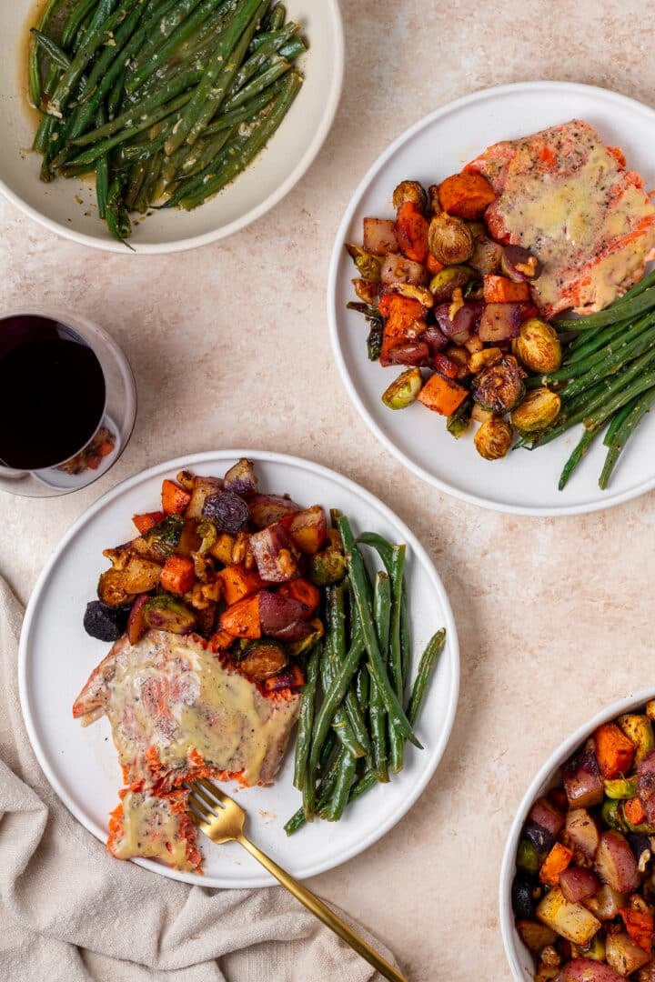 two plates with salmon and veggies, glass of red wine, gold fork, serving bowls of green beans and roasted vegetables