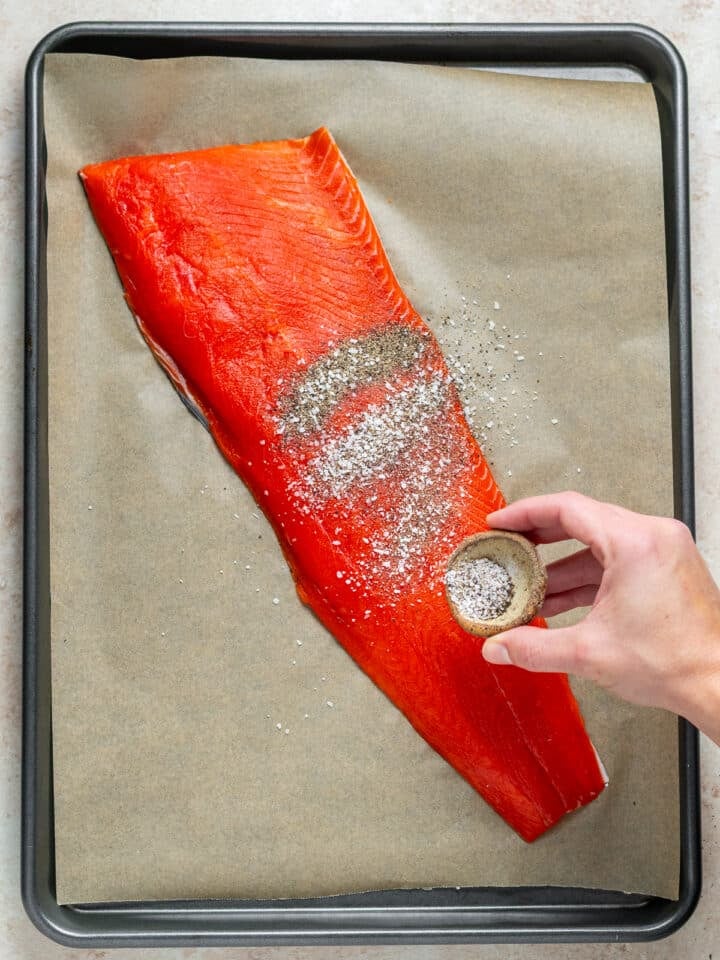 fillet of salmon on a sheet pan and a hand sprinkling it with salt and pepper