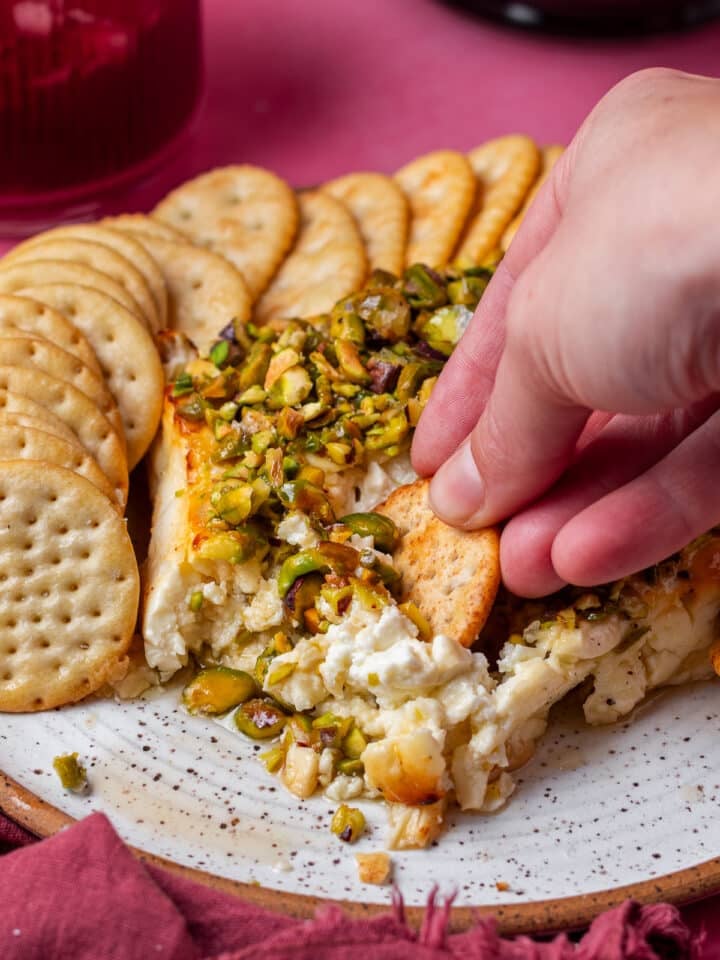 hand dunking a cracker into feta topped with pistachios and hot honey