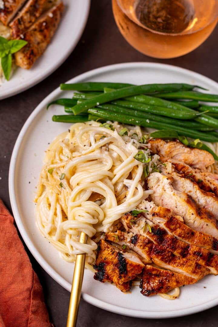 white plate with a gold fork twirling spaghetti alfredo, sliced grilled chicken and green beans