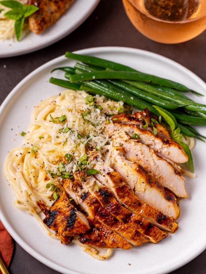spaghetti alfredo with sliced grilled chicken and green beans on a white plate, topped with basil and parmesan