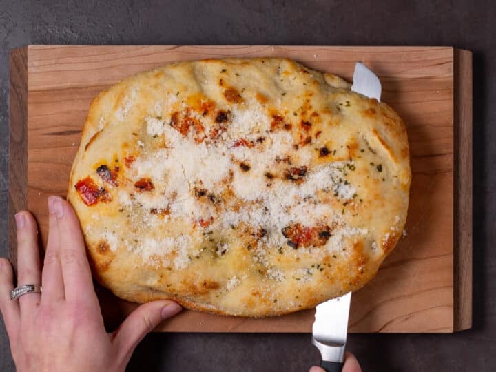 hands slicing a loaf of focaccia bread on a cutting board