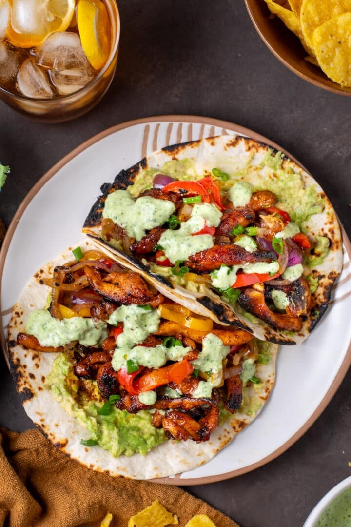 white plate with two fajitas filled with chipotle chicken, sauteed veggies, guacamole and cilantro lime yogurt sauce