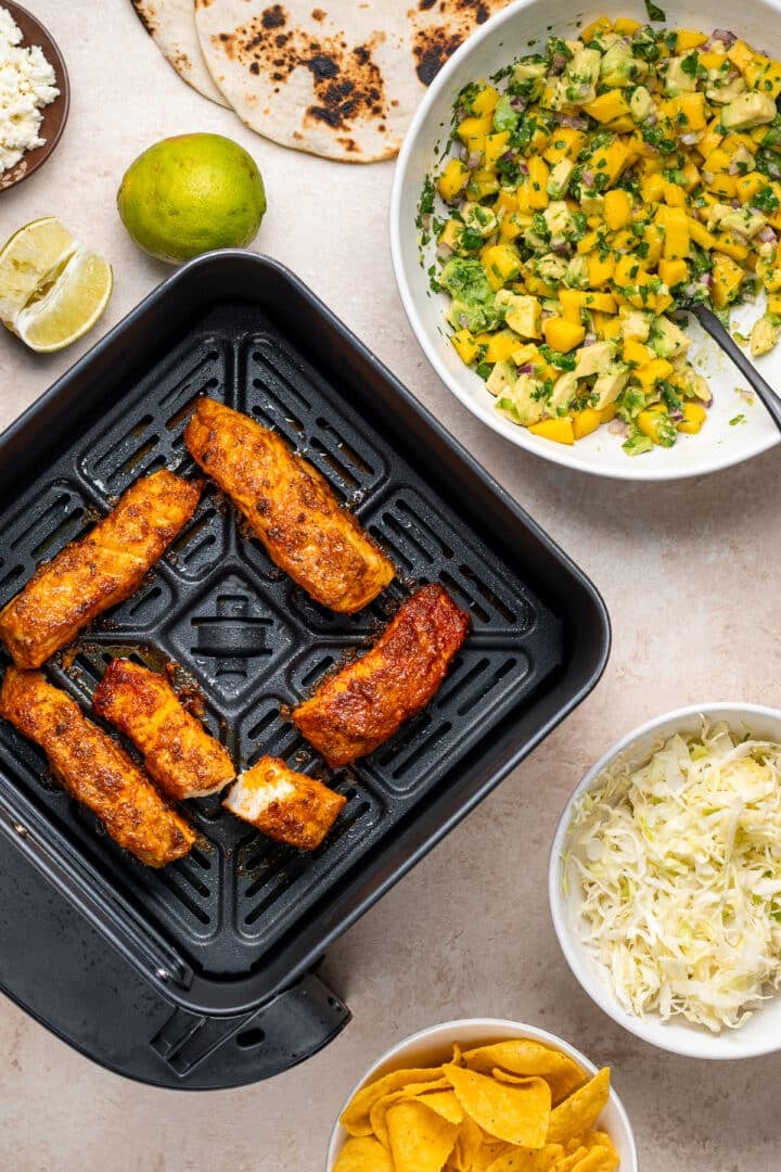 components of the air fryer fish tacos ready for assembly: fish in air fryer, mango pico de gallo, charred tortillas, cabbage and queso fresco