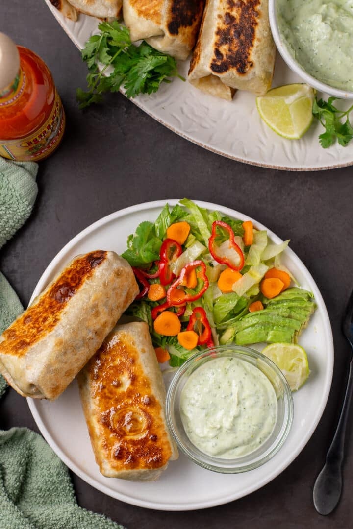 overhead view of two crispy burritos on a plate with a small bowl of creamy sauce and side salad
