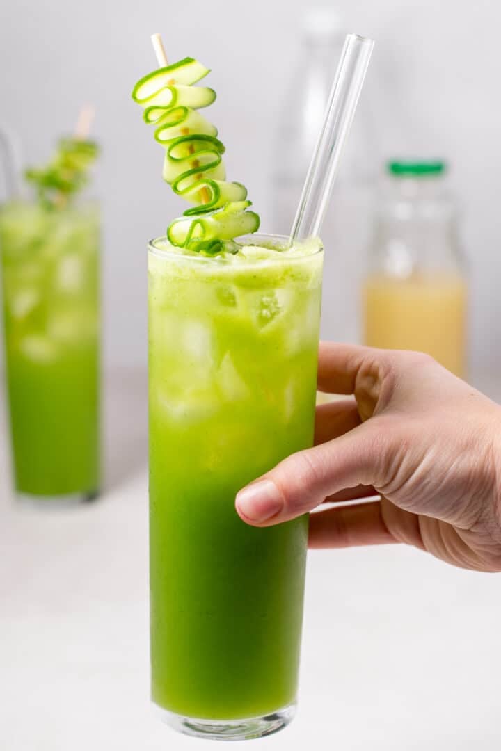 hand holding a glass of cucumber spritz garnished with a cucumber ribbon and glass straw