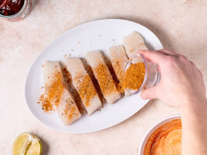 hand pouring spicing out of small glass bowl onto five fillets of white fish