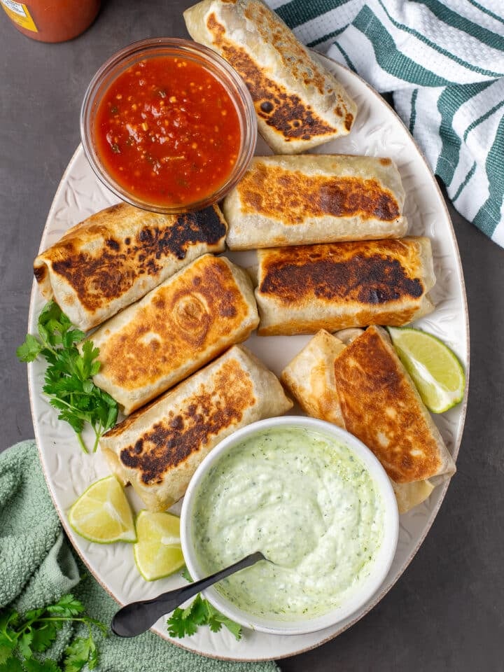 large white serving platter with cheesy bean and rice burritos, bowl of salsa and bowl of cilantro yogurt sauce