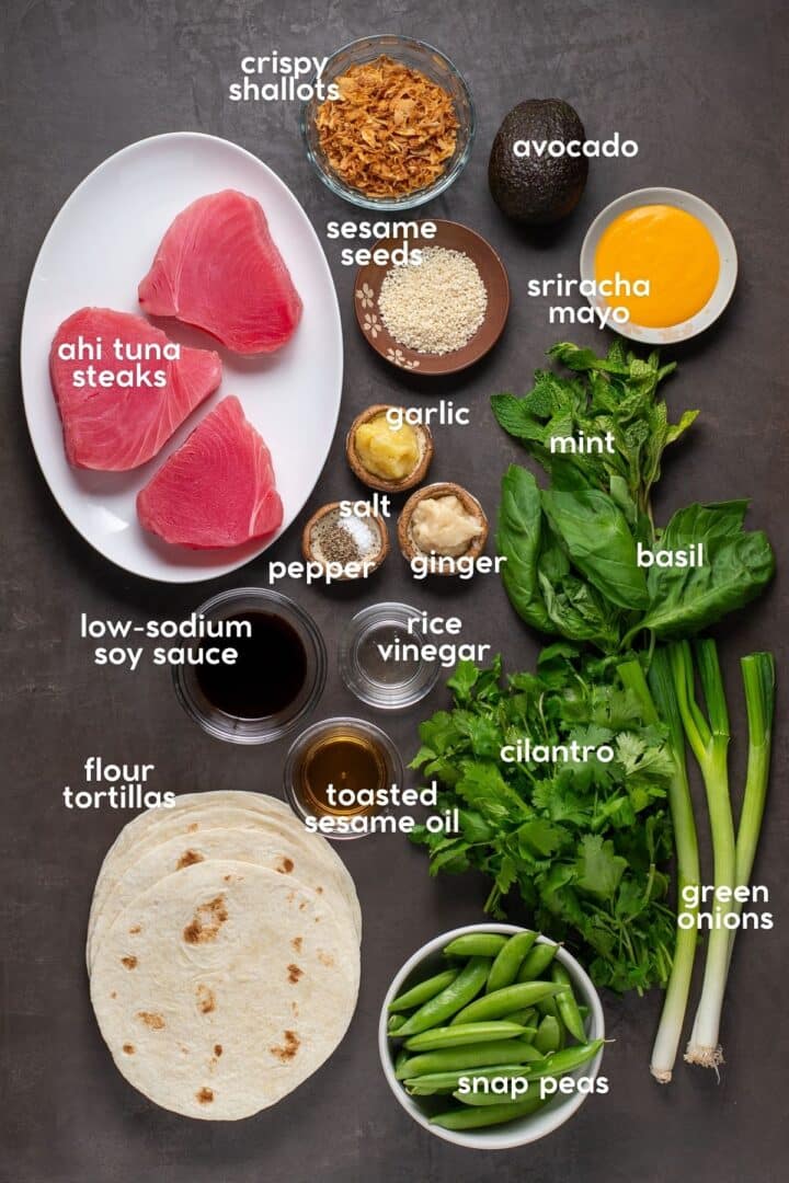 Ingredients for seared ahi tuna smash tacos including tuna steaks, snap peas, fresh herbs, flour tortillas, soy sauce, sesame oil, and sriracha mayo