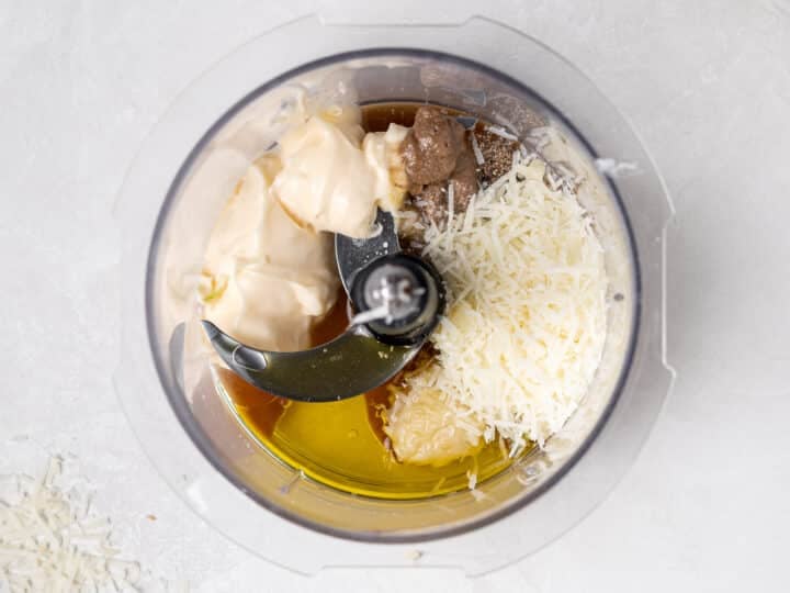 ingredients for Caesar vinaigrette in food processor including mayo, olive oil, parmesan, and anchovy paste