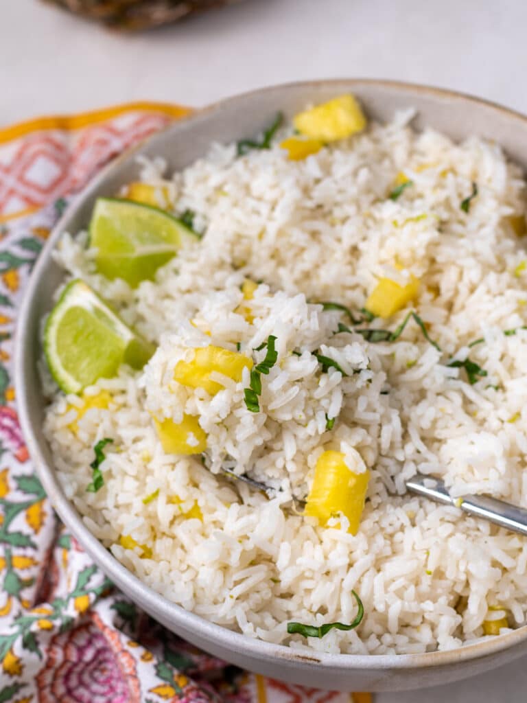 Bowl of pineapple coconut rice side dish with lime wedges, fresh herbs, and a fork scooping a bite