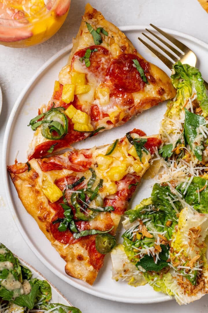 Close-up of pepperoni pineapple pizza slices on a plate with jalapeños, melted cheese, and Caesar salad with parmesan and crispy shallots