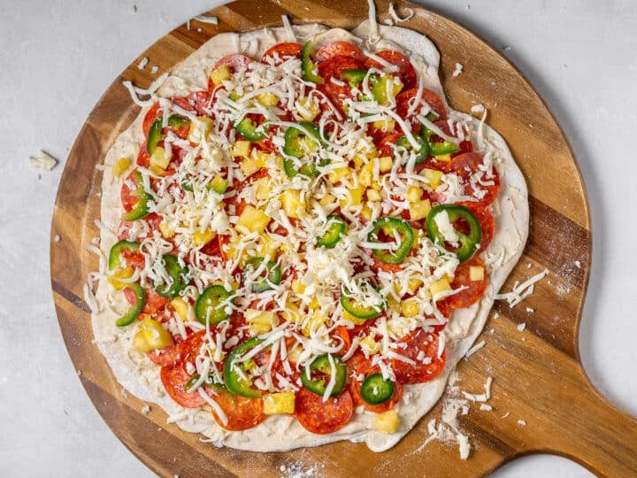 Unbaked pepperoni pineapple jalapeño pizza with creamy garlic sauce, layered with mozzarella cheese on pizza dough