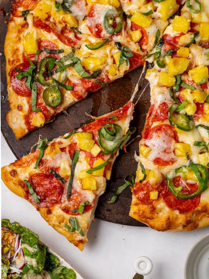 Pepperoni pineapple pizza with jalapeño slices and fresh basil, showing a slice being pulled away with melted mozzarella and diced pineapple